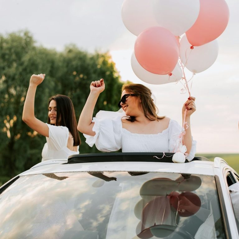 Two women celebrate in a car, holding pink and white balloons, with joyful expressions.